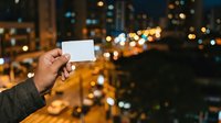 Hand holding blank white card against night cityscape with golden bokeh lights from urban skyline. Use for business, marketing, advertising, or promotional campaigns.