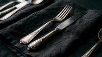 Vintage silver fork and knife with ornate handles arranged on dark black linen fabric, elegant table setting for fine dining, formal events, restaurants, hospitality design, and luxury tableware styling.