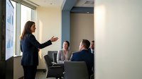 Professional businesswoman presenting to colleagues in modern conference room during corporate meeting with natural lighting from large windows.