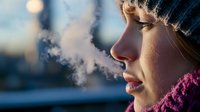 Young Asian woman in winter hat and scarf exhaling visible breath in cold winter air during sunset, side profile view capturing contemplative moment in frosty weather.