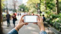 Person holding blank smartphone horizontally in urban park setting with blurred pedestrians and green trees in background for app mockup or mobile display advertising.