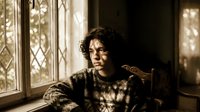 Young man with curly dark hair in patterned sweater sitting by ornate window with warm sunlight creating shadows in cozy indoor setting for lifestyle content.