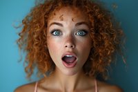 Young woman with bright blue eyes and natural curly red hair expressing surprise with open mouth against turquoise background, beauty portrait in natural light.