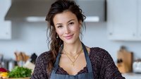 Young woman with long brown hair wearing purple cardigan and apron smiles in modern kitchen setting, creating warm homey atmosphere for cooking blogs and lifestyle content.