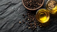 Black peppercorns scattered on dark stone surface with olive oil in glass bowls, overhead view showing texture and ingredients for cooking and seasoning.