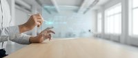 Male professional in business casual attire interacting with holographic digital interface on wooden table in modern office space with natural light and blurred background.