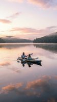 Couple kayaking on calm lake at sunrise with forested mountains and pink sky reflected in water, outdoor recreation and adventure.