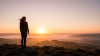 Woman with long dark hair standing on rocky mountain peak overlooking misty valley at sunrise, wearing casual dark clothing, contemplative pose for travel and wellness concepts.