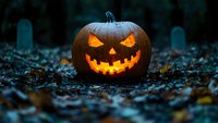 Glowing carved pumpkin with scary face in dark cemetery surrounded by fallen autumn leaves, creating spooky Halloween atmosphere at dusk.