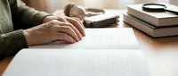 Senior person writing on paper at wooden desk with books and glasses nearby for education or business documentation.