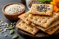 Crispy whole grain crackers with sunflower seeds and pumpkin seeds on wooden board, bowl of raw seeds and orange gerbera flower for healthy snacking.