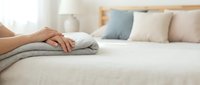 Hands placing folded gray towels on a neatly made bed in a bright, cozy bedroom with soft neutral tones and pastel pillows.