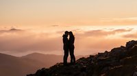 Romantic couple silhouette embracing on mountain peak during golden sunset with layered hills and warm sky creating intimate moment for love concepts.
