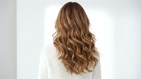 Young woman with long wavy brown hair with blonde highlights wearing white sweater from behind showing healthy hair styling and color treatment results.