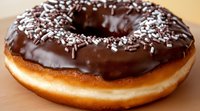 Fresh baked donut covered with dark chocolate glaze and white sprinkles on wooden surface, macro food photography showing texture and shine.