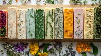Handmade soap bars with natural herbs, flowers and botanicals arranged in colorful rows showing variety of organic ingredients and textures on wooden surface.