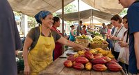 Smiling African American female vendor in yellow apron selling fresh colorful bell peppers at bustling outdoor farmers market under canvas tent with customers browsing.