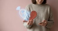 Young woman in beige sweater holding smartphone with glowing digital heart and wifi symbols representing online dating app connection and modern romance technology.