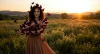 Young woman with autumn leaves crown in golden sunset field wearing flowing dress for seasonal portrait photography.