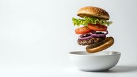 Floating hamburger with beef patty, lettuce, tomato and onion hovering above white bowl on clean background for food advertising and menu design.