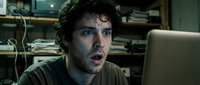 Young adult male with dark curly hair and light skin tone, appearing shocked or surprised while working on laptop in home office with shelving behind him.
