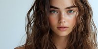 Young Caucasian woman with natural wavy brown hair and striking blue eyes against light background, bare shoulders, minimal makeup look showcasing freckles and glowing skin.