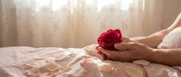 Romantic morning scene with hands holding red rose on bed with soft sunlight streaming through curtains creating intimate atmosphere for love concepts.