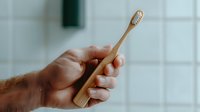 Male hand holding eco-friendly bamboo toothbrush against white tiled bathroom wall background, demonstrating sustainable oral hygiene and zero waste lifestyle choice.