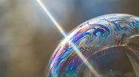 Iridescent soap bubble with rainbow swirls floating in sunlight creating prismatic reflections and colorful surface patterns against blurred background.