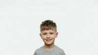 Portrait of a young boy with light brown hair and fair complexion, wearing a gray t-shirt, smiling at camera against neutral background for stock photography.