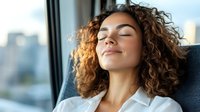 Young woman with curly hair relaxing in comfortable chair near window, eyes closed with peaceful smile, enjoying moment of tranquility in urban setting.
