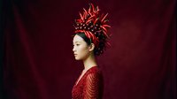 An Asian woman wearing an elaborate red feathered headdress and matching embroidered garment poses in profile against a deep burgundy backdrop, suitable for fashion, editorial, and cultural content.