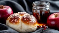 Fresh baked challah bread roll with sesame seeds on dark background with honey jar and red apples, traditional Jewish Shabbat or holiday food.