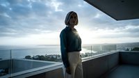 Professional businesswoman standing on modern balcony overlooking ocean view during golden hour sunset with dramatic cloudy sky and contemporary architecture.