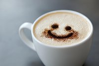 Creamy cappuccino in white ceramic cup decorated with cocoa powder smiley face pattern on milk foam, photographed against blurred gray background.