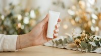 Person with medium skin tone holding blank white cosmetic tube against blurred botanical background with dried plants and natural lighting for skincare product mockup and beauty branding.