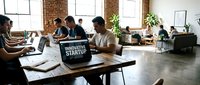 Diverse team of professionals collaborating in modern loft office space with laptops, notebooks, and innovative startup signage during brainstorming session.