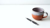 Orange and white ceramic coffee mug with black coffee next to black pen on clean white desk surface for morning productivity and workspace concepts.