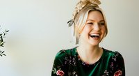 Young woman with blonde hair styled in an updo, fair complexion, wearing a dark green floral sweater, smiling brightly against a neutral background. Suitable for lifestyle, fashion, and portrait projects.