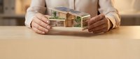 Hands protecting miniature modern house model with glass walls and green plants on wooden desk for real estate and property insurance concepts.