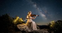 Young woman violinist in elegant gown performing under starry night sky with Milky Way galaxy visible above, creating magical outdoor concert atmosphere.