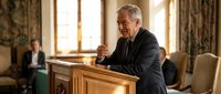 Senior Caucasian male priest delivering sermon at wooden pulpit in ornate church interior with congregation member seated in background pews.
