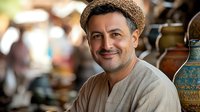 Middle-aged Mediterranean man with stubble wearing straw hat and linen shirt smiles warmly at camera, with decorative pottery and blurred market background.
