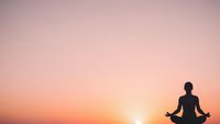 Woman silhouette meditating in lotus pose against warm sunset sky gradient background for wellness and mindfulness concepts.