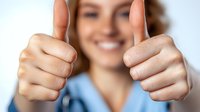 Young female healthcare worker in blue scrubs showing thumbs up gesture with both hands, expressing positivity and success in medical setting.