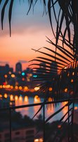 Palm fronds silhouetted against urban sunset skyline with bokeh city lights reflecting on water, creating dreamy tropical atmosphere in twilight hours.