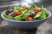 Fresh mixed green salad with sliced avocado, cherry tomatoes, and purple onion in blue ceramic bowl on rustic wooden table, healthy eating concept.