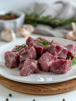 Fresh raw beef chunks cut for stew on white plate with rosemary sprigs, garlic cloves and peppercorns, served on wooden cutting board.
