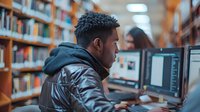 Young African American male student working on computer in university library, focused on digital screen with bookshelves in background.