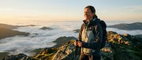 Middle-aged male hiker with glasses standing on mountain peak above clouds at sunrise, wearing outdoor gear and backpack for adventure and travel content.